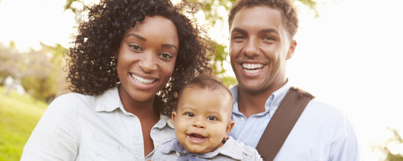 Family With Baby Son In Carrier Walking Through Park